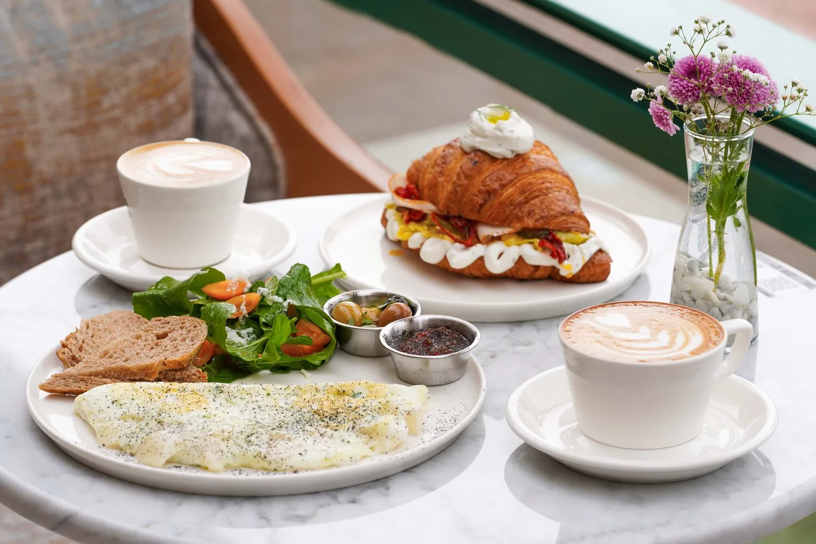 Brunch table spread with a croissant sandwich, two cups of coffee, an omelette, and a fresh salad